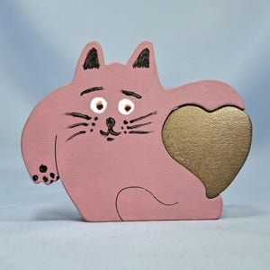 以下が含まれることがあります： 金色のハート型の切り抜きが付いた、ピンク色の木製の猫型フォトフレーム。猫には黒いひげ、目、耳のディテールがあります。フレームはマット仕上げで塗装されており、気まぐれで手作りの外観をしています。