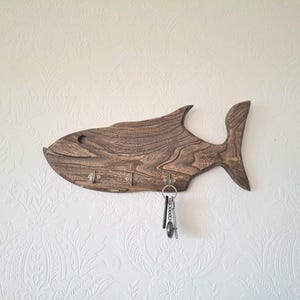 May include: A wooden fish-shaped key holder with a dark brown finish. The key holder has five metal hooks and a set of keys hanging from one of the hooks. The key holder is mounted on a white wall.