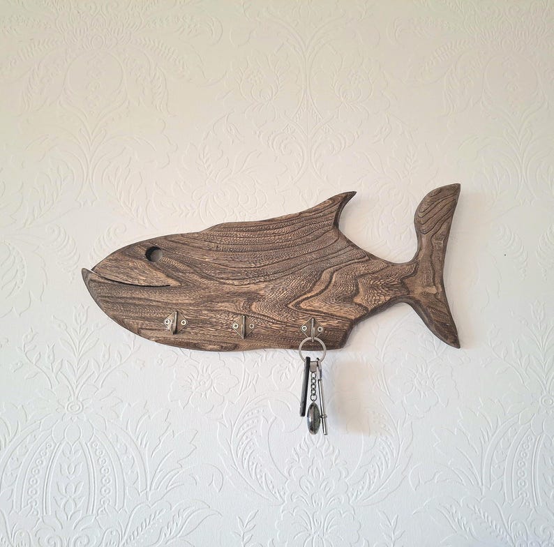 May include: A wooden fish-shaped key holder with a dark brown finish. The key holder has five metal hooks and a set of keys hanging from one of the hooks. The key holder is mounted on a white wall.