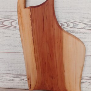 Puede incluir: Una tabla de servir de madera con borde natural y acabado liso y pulido. La tabla presenta un patr&oacute;n bicolor con un centro de color marr&oacute;n rojizo intenso y bordes m&aacute;s claros de color crema, incluyendo un asa integrada en la parte superior.