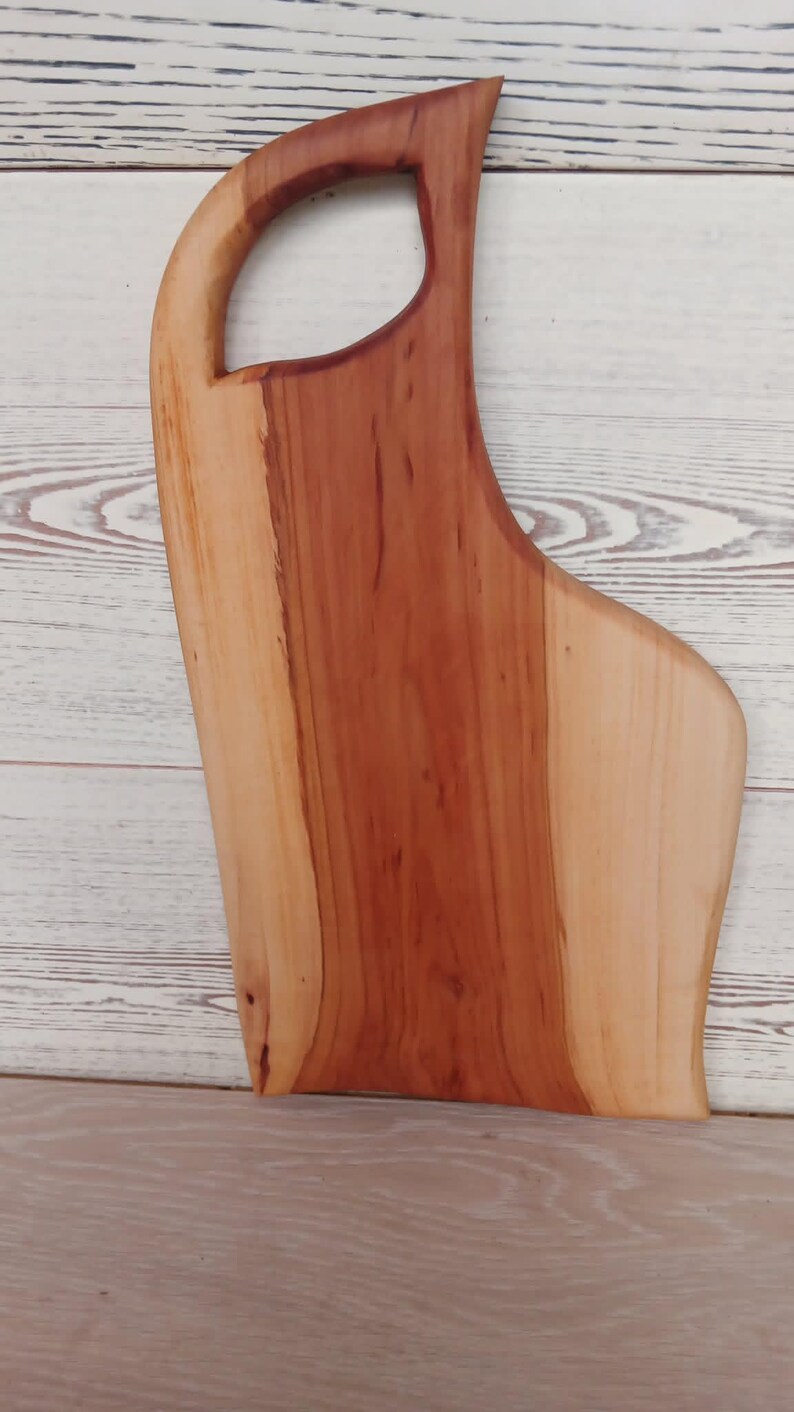 Puede incluir: Una tabla de servir de madera con borde natural y acabado liso y pulido. La tabla presenta un patr&oacute;n bicolor con un centro de color marr&oacute;n rojizo intenso y bordes m&aacute;s claros de color crema, incluyendo un asa integrada en la parte superior.