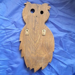 May include: Wooden owl-shaped wall decor with a natural wood grain finish. The owl has two circular eye holes and a small beak indentation. Two metal brackets are attached to the back for hanging. The owl's body is a textured, leaf-like shape.