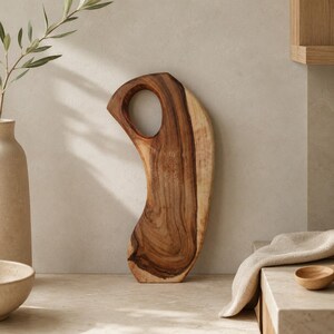 May include: A curved wooden cutting board with a natural grain pattern and a circular cutout handle at the top. The board features a deep brown centre with lighter wood tones along the edges, showcasing a smooth, organic shape and texture.