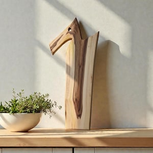May include: A tall, vertical wooden cutting board with a distinct bird-like silhouette, featuring a natural light and dark brown wood grain pattern and a circular cutout near the top. Beside it sits a white bowl filled with green herbs.