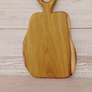 May include: A light brown wooden serving board with a pear-shaped silhouette and a smooth, polished finish. The board features a prominent vertical grain pattern and a circular cutout at the top of the handle for hanging or easy gripping.