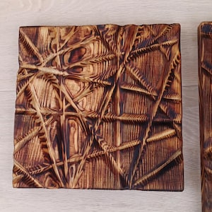 May include: Two square wooden panels with a geometric, textured design. The wood has a warm, brown tone with darker, burnt accents in the carved areas. The design features intersecting lines and shapes.