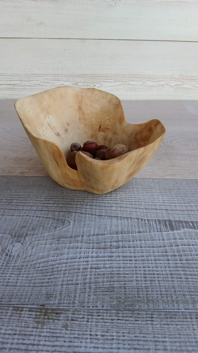 Op de afbeelding: Een handgemaakte houten kom met een natuurlijke, lichtbruine afwerking. De kom heeft een organische vorm en bevat verschillende noten. De kom wordt gepresenteerd op een houten oppervlak.