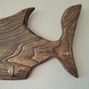 May include: A wooden fish-shaped wall hook with a dark brown finish. The hook has a textured surface and a bronze-colored metal hook. The design includes fins and a tail, adding a decorative element to the functional piece.