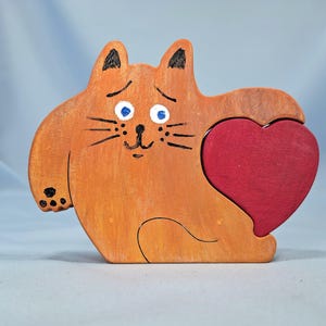 以下が含まれることがあります： 赤いハートを持つ木製のオレンジ色の猫の置物。猫は黒いひげ、青い目、耳と足に黒いアクセントがあります。ハートは濃い赤色です。