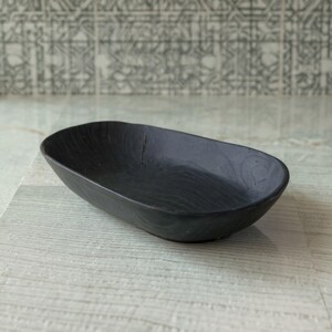 May include: A black, oval-shaped wooden bowl with a textured surface. The bowl has a smooth interior and a slightly raised rim. The dark color and simple design make it suitable for various uses, such as serving snacks or holding small items.