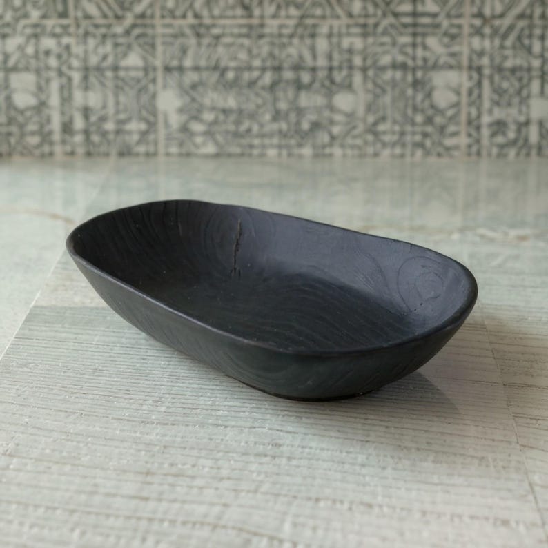 May include: A black, oval-shaped wooden bowl with a textured surface. The bowl has a smooth interior and a slightly raised rim. The dark color and simple design make it suitable for various uses, such as serving snacks or holding small items.