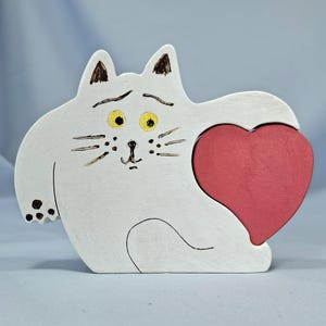 以下が含まれることがあります： 赤いハートを持つ白い木製の猫の置物。猫は黄色の目、黒いひげ、茶色のアクセントがあります。ハートは猫の腕の中にあります。猫はアンティーク風に塗装されています。