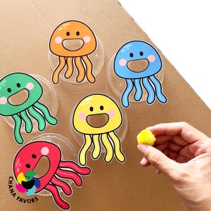 May include: Five colourful jellyfish with smiling faces are printed on white circles. The jellyfish are orange, blue, green, yellow, and red. A yellow pom-pom is in the foreground.