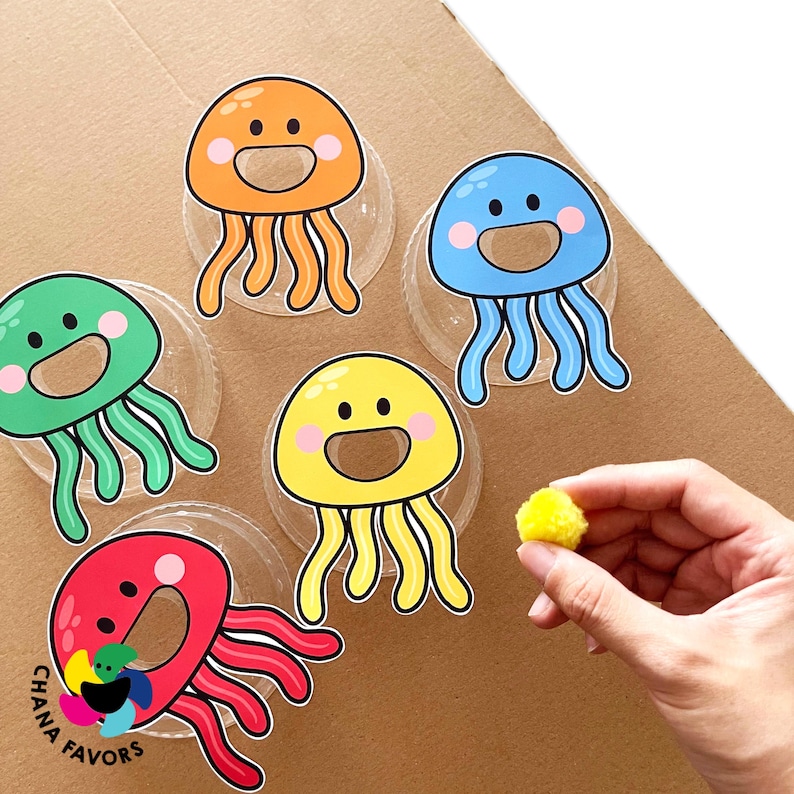 May include: Five colourful jellyfish with smiling faces are printed on white circles. The jellyfish are orange, blue, green, yellow, and red. A yellow pom-pom is in the foreground.