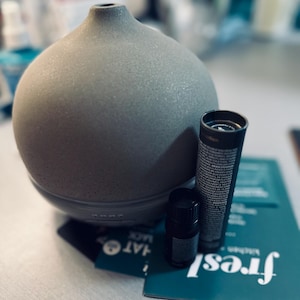 May include: A gray ceramic diffuser with a round base and a wide opening. The diffuser is sitting on a table with a small bottle of essential oil and a box with the word "Fresh" printed on it.