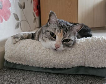 May include: A gray and white tabby cat with green eyes is lying on a white and gray fluffy bed. The cat is looking directly at the camera.