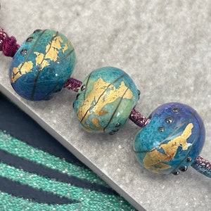 May include: Three glass beads with a blue, green, and purple gradient. Each bead has a gold leaf design that resembles a world map. The beads are strung on a purple ribbon.