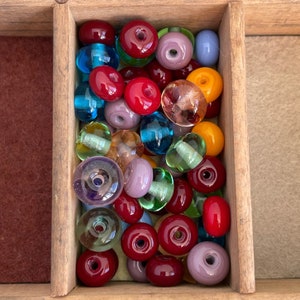 May include: A collection of colourful glass beads in various shapes and sizes, including round, oval, and teardrop. The beads are arranged in a wooden box, with some beads overlapping.