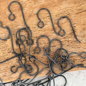 May include: Close-up of several dark gray metal earring hooks. The hooks are in various positions, some overlapping, against a light brown wooden surface. The hooks have a simple, curved design with a small loop at the end.