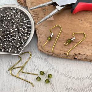 May include: A close-up of earring making supplies, including gold earring hooks, green glass beads, and silver beads. The supplies are arranged on a wooden surface.