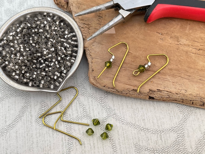 May include: A close-up of earring making supplies, including gold earring hooks, green glass beads, and silver beads. The supplies are arranged on a wooden surface.