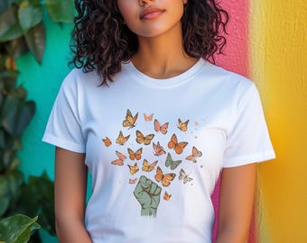 May include: White t-shirt featuring a graphic of a green fist releasing a swarm of orange and pink butterflies. The butterflies are in various sizes and designs. The background is a colorful wall.