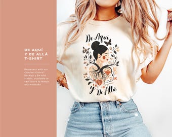 May include: A white t-shirt with a graphic design of a woman with a tree growing from her head. The text "De Aqui y De Alla" is printed on the shirt.