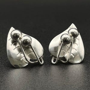 May include: A pair of silver-toned earrings shaped like leaves with two small, polished spherical beads attached to the front. The leaf design features etched lines for texture, and the metal has a reflective, smooth finish throughout the piece.