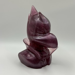 May include: A small, purple figurine of a creature with a pointed head and wings. The figurine is made of resin and has a glittery finish.