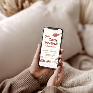 Może przedstawiać: Smartfon wyświetla zaproszenie urodzinowe z napisem "Our Little Meatball is turning one!" w czerwonym skrypcie. Zaproszenie zawiera ilustracje makaronu i czosnku. Telefon jest trzymany w dłoniach osoby.