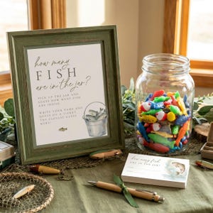 Puede incluir: Un letrero enmarcado con el texto "How many fish are in the jar?" está junto a un frasco de vidrio lleno de peces de juguete coloridos. También hay un bolígrafo y tarjetas con el mismo texto, junto con señuelos de pesca y una red.