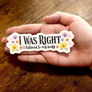 Könnte beinhalten: Ein weißer Aufkleber mit dem Text "I WAS RIGHT I listened to my body" in Schwarz. Der Aufkleber ist mit kleinen lila, gelben und rosa Blumen verziert. Der Aufkleber wird in einer Hand gehalten.