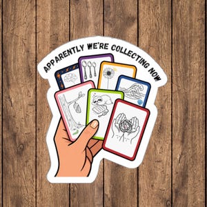 Peut inclure: Un autocollant représentant une main tenant un éventail de cartes colorées avec diverses illustrations au trait, dont une chrysalide, un paresseux et des mains tenant un enchevêtrement. Le texte "APPARENTLY WE'RE COLLECTING NOW" est en noir.
