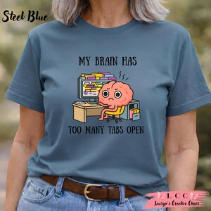 K&ouml;nnte beinhalten: Stahlblaues T-Shirt mit einer humorvollen Grafik. Das Design zeigt ein Cartoon-Gehirn, das an einem Schreibtisch vor einem Computer sitzt, mit dem Text "MY BRAIN HAS TOO MANY TABS OPEN".