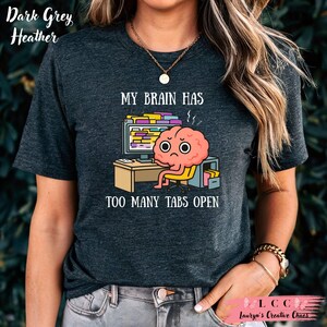 K&ouml;nnte beinhalten: Dunkelgraues Heather-T-Shirt mit dem Text "MY BRAIN HAS TOO MANY TABS OPEN". Die Grafik zeigt ein Cartoon-Gehirn, das an einem Schreibtisch vor einem Computer mit vielen offenen Tabs sitzt.