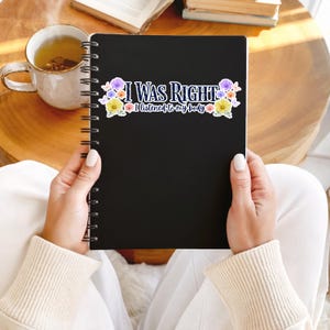 May include: A black spiral-bound notebook held by a person. The notebook has the text "I WAS RIGHT I listened to my body" with floral accents. A cup of tea and a book are in the background.