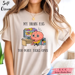 May include: Soft cream-colored t-shirt featuring the text "MY BRAIN HAS TOO MANY TASKS OPEN" above a cartoon brain character sitting at a desk in front of a computer. The brain has a stressed expression.