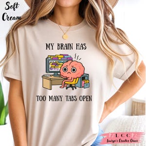 K&ouml;nnte beinhalten: Cremefarbenes T-Shirt mit dem Text "MY BRAIN HAS TOO MANY TABS OPEN". Die Grafik zeigt ein Cartoon-Gehirn, das an einem Schreibtisch vor einem Computer sitzt. Das Gehirn hat einen gestressten Ausdruck. Das Shirt hat auch den Text "Lauryn's Creative Chaos."
