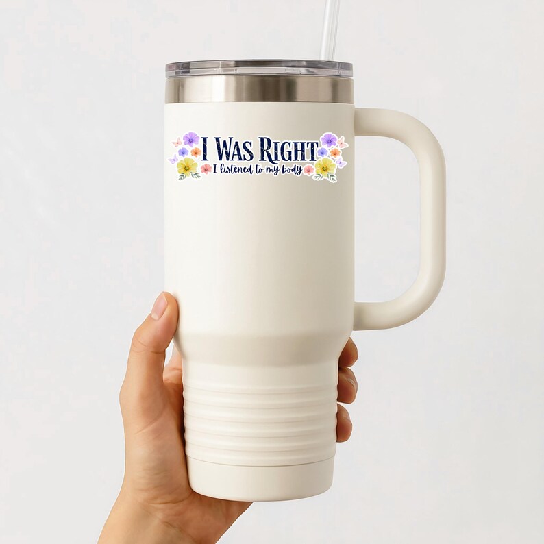 May include: A white travel mug with a silver lid and a handle. The mug has the text "I WAS RIGHT" with the phrase "I listened to my body" below it, and is decorated with floral accents. A straw is inserted into the lid.