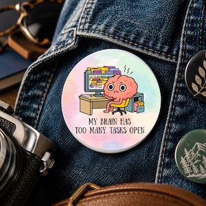 May include: A round pinback button featuring a pastel watercolor background and a cartoon illustration of a pink brain sitting at a desk with a computer. The text reads: MY BRAIN HAS TOO MANY TASKS OPEN. The button is attached to a blue denim fabric.