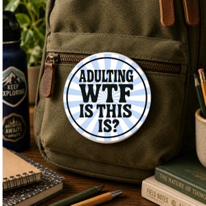 May include: A circular pinback button featuring a light blue and white sunburst pattern with the black text ADULTING WTF IS THIS IS? centered on the surface. The button is attached to the front pocket of an olive green canvas backpack.