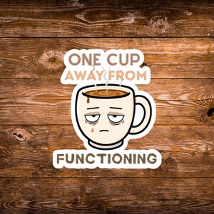 May include: A die-cut sticker featuring a cartoon coffee mug with a tired, sleepy face and dark circles under its eyes. The mug contains brown liquid with steam rising above it. The text reads ONE CUP AWAY FROM FUNCTIONING in brown letters.