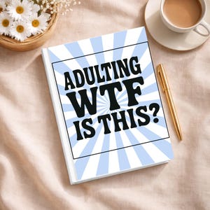 May include: A rectangular notebook with a white cover featuring a light blue sunburst pattern and the black text ADULTING WTF IS THIS? centered in a black-bordered box. A gold pen is positioned to the right of the notebook on a light fabric.
