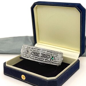 May include: A silver-toned filigree cuff bracelet featuring an intricate openwork metal design with small embedded clear stones and a single square-cut green stone, presented inside a navy blue velvet-lined jewelry box with a cream interior.
