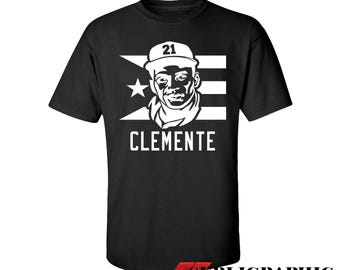 May include: Black t-shirt featuring a white graphic of a baseball player wearing a cap with the number 21. The graphic includes the Puerto Rican flag and the name "CLEMENTE" in bold letters.