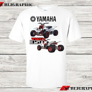 Könnte beinhalten: Weißes T-Shirt mit einer Grafik von zwei Yamaha ATVs mit roten und weißen Akzenten. Das Yamaha-Logo und der Text "TWIN 350" sind über und unter den ATV-Illustrationen aufgedruckt. Das Shirt hat einen Rundhalsausschnitt.