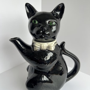 May include: A black ceramic cat-shaped teapot with green eyes and a cream bow tie. The teapot has a spout and handle. The glossy finish reflects light. The teapot is approximately 25 cm tall, designed to sit upright.