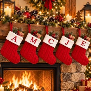 May include: Five red cable-knit Christmas stockings with white cuffs, each featuring a single embroidered red letter: A, M, C, J, and E. Each stocking is filled with pine sprigs, red berries, and a small gift box tied with twine.