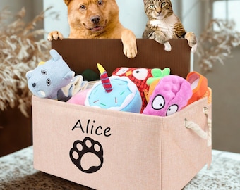 May include: A beige fabric pet toy storage bin with the name "Alice" and a paw print. The bin is filled with colorful plush toys, and a dog and cat are peering over the top of a brown box.