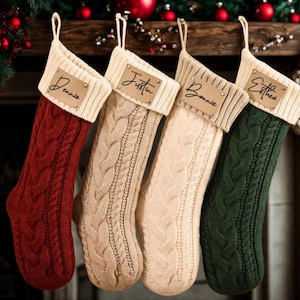 Könnte beinhalten: Vier Zopfstrick-Weihnachtsstrümpfe in Rot, Beige, Creme und Grün, jeweils mit cremefarbenem Bündchen und einem Namensschild aus Leder. Die Namen "Dennis", "Justin", "Bonnie" und "Esther" sind auf den Etiketten abgebildet. Die Strümpfe hängen an einem Holzkamin.
